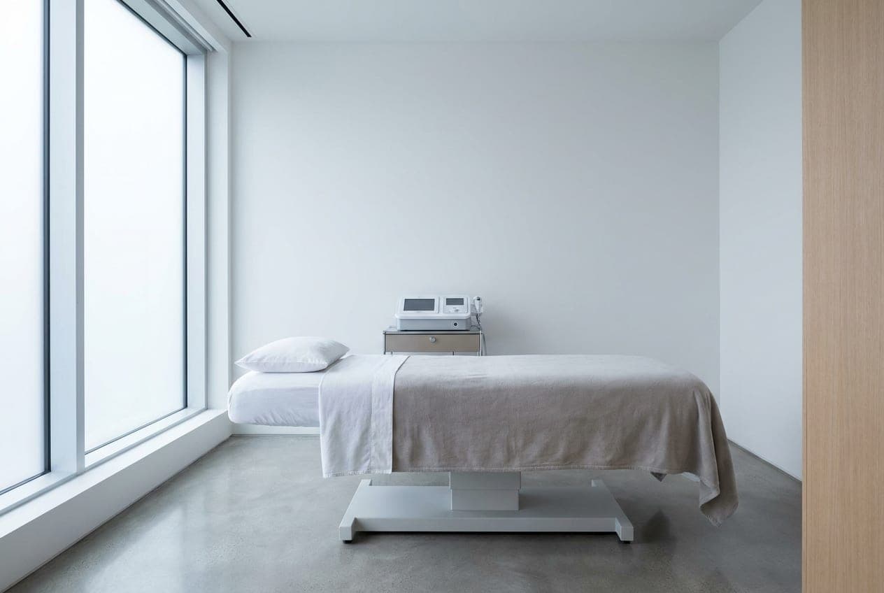 The Lumera body contouring room — a treatment bed in cool linen beside a low equipment cart